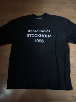 Svart Acne Studios t-shirt - Säljer en svart t-shirt från Acne Studios med vit text på bröstet där det står 'Acne Studios STOCKHOLM 1996'. Klassisk passform och rund halsringning. Perfekt till jeans eller shorts för en clean och stilren look.