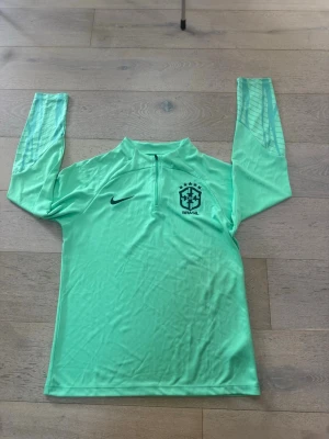 Mintgrön Brasilien träningströja Nike - Snygg mintgrön långärmad träningströja från Nike med Brasilien-logga och halv dragkedja framtill. Tröjan har diskreta ränder på ärmarna och är tillverkad i ett lätt, funktionsmaterial som passar perfekt till fotbollsträning eller annan sport.