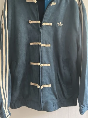 Blå adidas jacka, chinese new year edition - Unik blå adidas jacka med beige snörningsdetaljer och klassiska tre ränder längs ärmarna. Jackan har dragkedja framtill, elastisk nederkant och adidas-logga på bröstet. Perfekt för dig som vill sticka ut med sportig stil.
