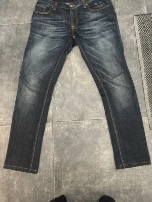 Mörkblå jeans från Nudie Jeans - Säljer ett par mörkblå jeans från Nudie Jeans med snygga slitningar och kontrastsömmar. Klassisk femficksmodell i rak passform och tillverkade i denim. Perfekta för dig som gillar stilrena och tidlösa jeans.