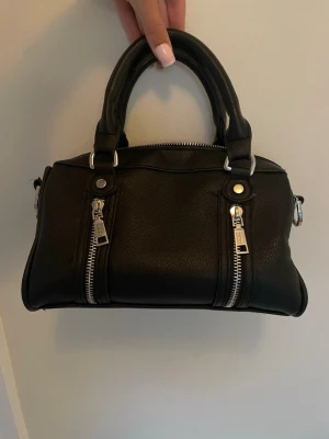Svart handväska med silverdragkedjor - Snygg svart handväska i fejkläder med två korta handtag och coola silverfärgade dragkedjor fram och baktill. Köpt på Zalando och märket heter Misako. 