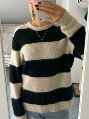 Randig stickad tröja i svart och beige - Mysig stickad tröja med breda svarta och beige ränder. Tröjan har rund halsringning och långa ärmar. Perfekt för dig som gillar en avslappnad och trendig stil. Passar till både jeans och kjol. 10% ull, 4% alpacka.