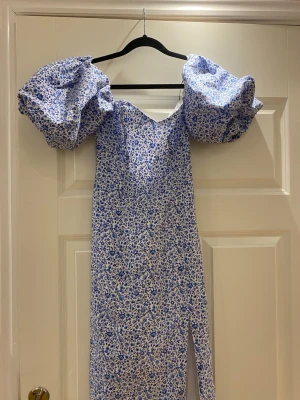 Blommig klänning med puffärmar - Supersöt lång klänning med blått och vitt blommönster och stora puffärmar. Klänningen har en slits framtill och snörning i ryggen för en snygg passform. Materialet ser ut att vara bomull eller bomullsblandning. Perfekt för dig som gillar statement-ärmar och romantisk stil.