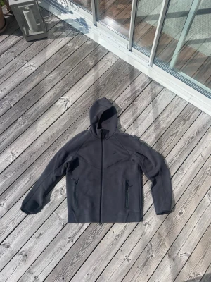NIKE tech fleece  - Säljer en stilren mörkgrå hoodie med hel dragkedja framtill och två fickor med dragkedja. Hoodien har en klassisk huva och är tillverkad i ett mjukt, tjockare material som passar perfekt för kyligare dagar. Enkel och clean design utan synligt märke.