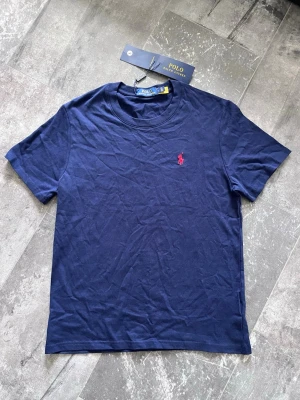 Polo Ralph Lauren T-shirt  - Helt ny klassiks marinblå t-shirt från Polo Ralph Lauren med korta ärmar och rund hals. Tillverkad i mjuk bomull och har den ikoniska röda logobrodyrn på bröstet. Enkel och stilren modell som funkar till det mesta.