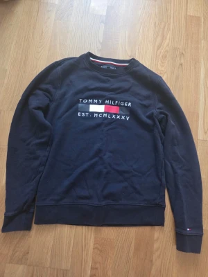 Mörkblå sweatshirt Tommy Hilfiger - Mörkblå sweatshirt från Tommy Hilfiger med klassisk logga och texten 'EST. MCMLXXXV' på bröstet. Tröjan har rund hals, långa ärmar och ribbade muddar. Perfekt för en avslappnad och stilren look.