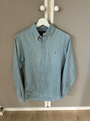 Ljusblå skjorta från Polo Ralph Lauren - Klassisk ljusblå skjorta från Polo Ralph Lauren i slim fit-modell. Skjortan har button-down krage, långa ärmar och den ikoniska broderade loggan på bröstet. Tillverkad i mjuk bomull och stängs med knappar framtill.