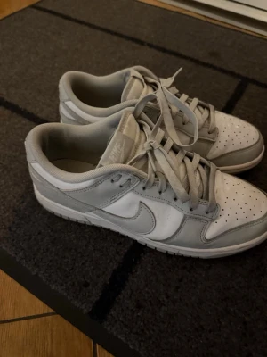 Nike vita och grå låga sneakers - Snygga sneakers från Nike i vitt och ljusgrått. Skorna har klassisk låg siluett, perforerad tåbox och ikoniska swoosh-loggan på sidan. Tillverkade i läder och textil med matchande vita och grå skosnören. Perfekta för en clean och stilren look.