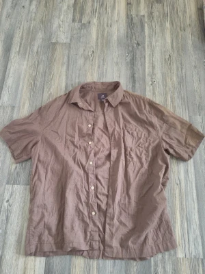Brun kortärmad skjorta från H&M - Säljer en brun kortärmad skjorta från H&M i regular fit. Skjortan har klassisk krage och knappar framtill. Tillverkad i ett lätt bomullsmaterial som passar perfekt till varmare dagar. Har en fläck på vänsterarm men den går bort i en maskintvätt.
