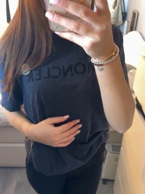 Svart Moncler t-shirt med logga - Snygg svart t-shirt från Moncler med klassisk logga på bröstet och tryckt Moncler-text. T-shirten har en avslappnad passform och är tillverkad i mjuk bomull. Perfekt för en stilren och trendig look.