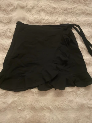 Svart omlottkjol med volang - Snygg svart omlottkjol med volangkant och knytband i midjan. Kjolen har en kort längd och är perfekt för dig som gillar en enkel men trendig stil. Passar till många olika outfits och är lätt att matcha.