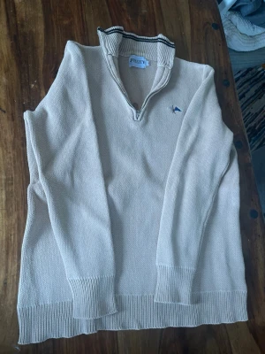 Beige stickad tröja från Jem City - Beige stickad tröja från Jem City med quarter zip dragkedja och ståkrage med svarta ränder. Tröjan har ribbstickade muddar vid ärmslut och nederkant samt en liten broderad logga på bröstet. Perfekt för lager på lager och enkel att matcha.