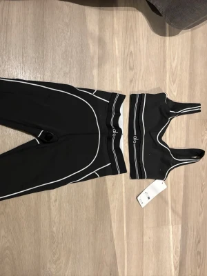 Svart träningsset från Alo Yoga - Snyggt svart träningsset från Alo Yoga med vita kontrastsömmar. Setet består av sport-bh med breda axelband och matchande tights med hög midja. Perfekt för gymmet eller yoga. Materialet är stretchigt och följsamt för maximal komfort.
