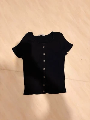 Svart T-shirt från H&M med knappar - Säljer en söt svart T-shirt med bruna knappar. Står strl 146/152 men tycker de passar bra Xs/ XXS då den är stretchig☺️