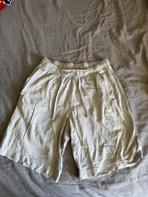 Gråa shorts från Nike med snörning - Säljer ett par ljusgrå shorts från Nike med elastisk midja och snörning. Shortsen har fickor på sidorna och en broderad Nike-logga på vänster ben. Perfekta för sommaren och riktigt sköna att ha på sig.