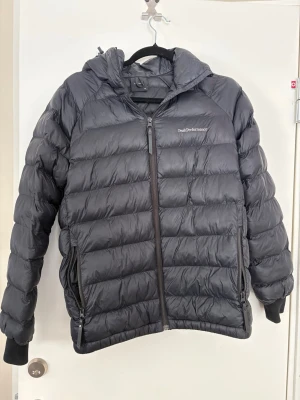 Svart dunjacka från Peak Performance - Snygg svart dunjacka från Peak Performance med huva och dragkedja framtill. Jackan har två sidofickor med dragkedja och ribbade muddar vid ärmsluten. Perfekt för kalla vinterdagar och riktigt skön att bära. Jackan har även ett litet hål i ena fickan och de kan göra att dragkedjan fastnar men de går enkelt att fixa. Storleken är 170 men passar en S/M också