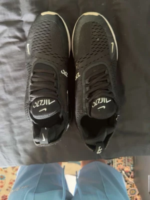 Nike Air Max 270 svarta sneakers - Svarta Nike Air Max 270 sneakers med vit logga och detaljer. Ovandelen är i mesh för bra ventilation och sulan har en tydlig synlig Air-enhet i hälen. Skorna har snörning och en sportig, modern look med inslag av vitt och rött på sulan.
