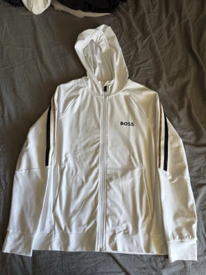 Vit hoodie med dragkedja från BOSS - Snygg vit hoodie från BOSS med dragkedja framtill och svarta ränder längs ärmarna. Klassisk huva och diskret BOSS-logga på bröstet. Tillverkad i ett mjukt material som känns skönt mot huden. Perfekt för en clean och sportig look.