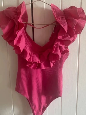 Rosa body med volang från Zara - Supersnygg rosa body från Zara med djup ringning och stora volangdetaljer runt halsen. Tillverkad i ett stretchigt, ribbat material som sitter tajt och bekvämt. 