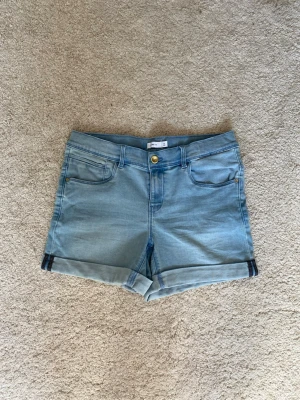 Ljusblå denimshorts - Säljer ett par ljusblå denimshorts med klassisk femficksdesign och uppvikta benslut. Shortsen har normal passform, bälteshällor och stängs med knapp och dragkedja. Bakfickorna har en snygg design. 