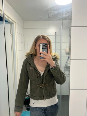 Vintage Hollister grön parkajacka med fuskpäls - Snygg vintage y2k grön parkajacka med huva och fuskpälsdetalj från Hollister & Co. Jackan har träknappar och snörning framtill samt en broderad logga på bröstet. 💗Vintage storlek L passar mer som M jag har S och kan ha en hoodie under. 💗Lite fransning runt manschetterna men inget som påverkar slitaget. 💗Fler bilder finns. 💗Ungefärliga platta mått: axel till axel: 36 cm axel till botten: 50 cm armhåla till armhåla: 46 cm yttre ärm: 63 cm innerarm: 47 cm