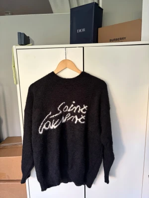 Svart stickad tröja Saint Laurent - Säljer en svart stickad tröja från Saint Laurent med vit logga i graffiti-stil på bröstet. Tröjan har rund halsringning och långa ärmar. Materialet ser mjukt och fluffigt ut, perfekt för kyligare dagar.