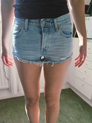 Ljusblå högmidjade jeansshorts W27 - Säljer ett par ljusblå högmidjade jeansshorts med råa, fransiga kanter och klassisk femficksdesign. Shortsen har en knappgylf och bälteshällor, perfekt för sommarens alla äventyr. Snygg vintagekänsla och avslappnad passform.