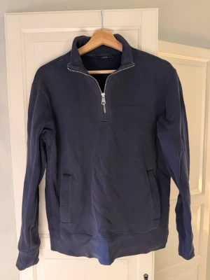 J.Lindeberg halfzip - Snygg marinblå tröja från J.Lindeberg med halv dragkedja och broderad logga på bröstet. Tröjan har en stor magficka framtill och är tillverkad i mjukt bomullsmaterial. Perfekt för en avslappnad men stilren look.