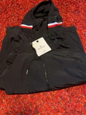 Svart vindjacka med huva från Moncler - Snygg svart vindjacka från Moncler med dragkedja framtill och huva. Jackan har en sportig look med röd, vit och blå rand på huvan samt justerbara snören. Tillverkad i lätt och tåligt material, perfekt för blåsiga dagar.