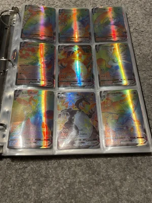 Pokémon samlarkort VMAX i pärm - Säljer en sida med Pokémon samlarkort, flera av dem är holografiska och har färgglada regnbågsmönster. Korten ligger i plastfickor och har klassisk blå baksida med Pokéball. Perfekt för dig som vill utöka din samling! Det är bara v max kort. Några är väldigt rare och kan kosta upp till 2000 kr