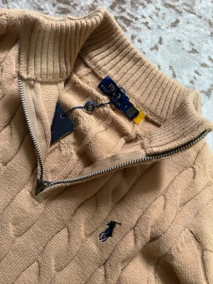 Polo Ralph Lauren - Snygg beige kabelstickad tröja från Polo Ralph Lauren med halv dragkedja och hög krage. Tröjan har ribbade muddar och nederkant samt den klassiska mörkblå loggan broderad på bröstet. Perfekt för en stilren och avslappnad look.