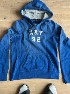 Blå hoodie med A&F 92 tryck - Snygg blå hoodie med huva och magficka. Framsidan har broderat A&F 92 i vitt. Insidan av huvan är ljusgrå med ribbad struktur. Hoodien har dragsko i huvan och är perfekt för en avslappnad stil.