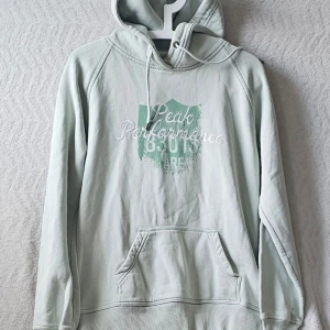 Ljusgrå hoodie Peak Performance - Snygg hoodie från Peak Performance med tryck och broderad text på bröstet. Klassisk känguruficka framtill och justerbar huva med dragsko. Perfekt för en avslappnad och sportig stil.