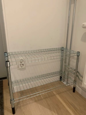 Metallhylla med två plan - Praktisk hylla i silverfärgad metall med två gallerplan och svarta plastfötter. Perfekt för förvaring av skor, väskor eller andra prylar i hallen. Som ny