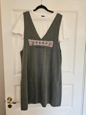Grå klänning med text från Gina Tricot - Grå ärmlös klänning från Gina Tricot med djup v-ringning och vit text 'WEEKEND EVERYDAY' framtill och vit tshirt fastsydd. Klänningen har en avslappnad passform och är gjord i mjukt material. Perfekt att styla över en t-shirt för en casual look.  Kan även hämtas I Partille  E4