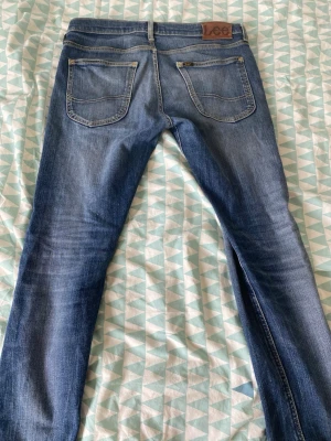 Blå jeans från Lee med smal passform - Säljer ett par klassiska blå jeans från Lee med smal passform och snygga slitningar. Jeansen har fem fickor, dragkedjegylf och Lee-lapp bak i midjan. Perfekta för en avslappnad och trendig look.