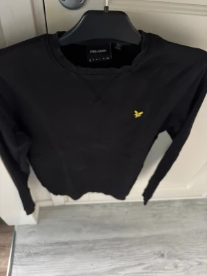 Svart sweatshirt från Lyle & Scott - Klassisk svart sweatshirt från Lyle & Scott med rund halsringning och gul broderad logga på bröstet. Tröjan har ribbade muddar vid ärmslut och nederkant samt en diskret detaljsöm vid halsen. Perfekt för en avslappnad stil.