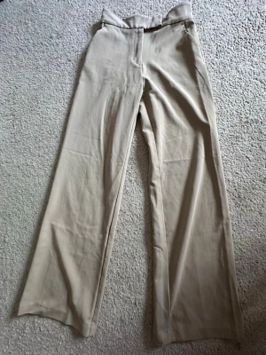 Beige kostymbyxor - Beige kostymbyxor med hög midja och rakvida ben. Byxorna har inga synliga fickor och är tillverkade i ett mjukt, lätt tyg. För långa för mig. Använda 2 gånger. 