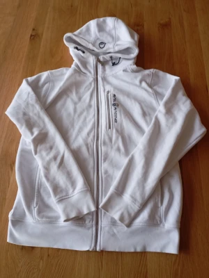 Vit sail racing kofta - Snygg vit hoodie från Sail Racing med dragkedja framtill och en extra bröstficka med dragkedja. Broderad logga på bröstet och huva med dragsko. Tillverkad i mjukt material som känns skönt mot huden. Perfekt för en avslappnad och sportig stil.