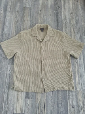 Grågrön kortärmad skjorta från H&M - Säljer en grågrön kortärmad skjorta från H&M i loose fit. Skjortan har knappar framtill, klassisk krage och är tillverkad i ett luftigt, ribbat material som passar perfekt till varmare dagar.