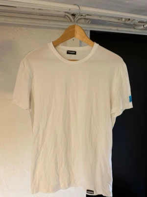 Vit t-shirt från Dsquared2 ICON - Säljer en clean vit t-shirt från Dsquared2 ICON med en blå patch på ärmen där det står 'ICON'. T-shirten har rund hals och är gjord i mjukt bomullstyg. Perfekt för dig som gillar stilrena plagg med en twist.