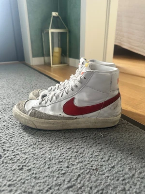 Nike vita high top sneakers röd swoosh - Klassiska vita high top sneakers från Nike med röd swoosh på sidan och Nike-logga bak. Skorna har vit ovandel i läder, grå mockadetaljer vid tån och vit sula. Perfekta för dig som gillar retrostil och streetwear. Funkar även bra att ha på gymmet!