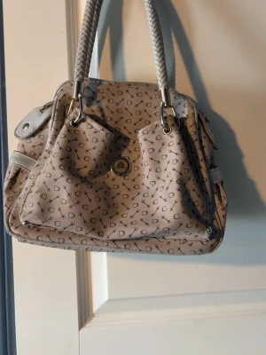 Beige handväska med mönster - Snygg beige handväska med diskret mönster i detaljer. Väskan har två flätade handtag, flera fack och guldfärgade metalldetaljer. Köpte på secondhand för ca 150kr, lite oklart vart det är ifrån 