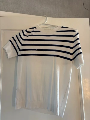 Vit topp med marinblå ränder - Vit topp med korta ärmar och marinblå horisontella ränder över bröst och axlar. Rund halsringning och ribbade kanter vid ärmslut och nederkant. Perfekt för en fräsch och stilren look.