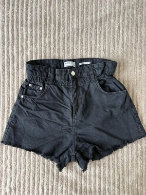 Svarta högmidjade shorts med rå kant - Säljer ett par svarta högmidjade shorts med rå, fransig kant och klassisk femficksdesign. Shortsen har knapp och dragkedja framtill samt en papperspåse-liknande midja för extra stilpoäng. Perfekta till sommaren!