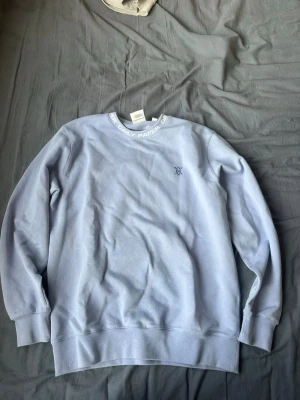 Ljusblå sweatshirt från Daily Paper - Ljusblå sweatshirt från Daily Paper med rund halsringning där märkesnamnet står tryckt. Tröjan har långa ärmar, ribbade muddar och en liten broderad logga på bröstet. Perfekt för en clean och avslappnad stil.