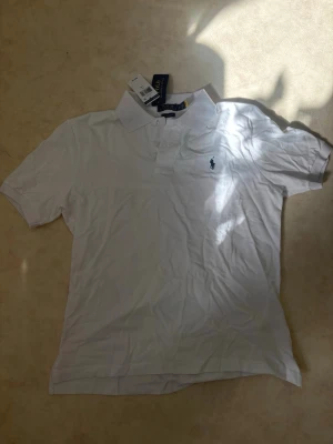 Vit pikétröja från Ralph Lauren - Klassisk vit pikétröja från Ralph Lauren med korta ärmar och den ikoniska broderade loggan på bröstet. Tröjan har krage och knappar framtill, perfekt för en clean och stilren look. Storlek L slim fit