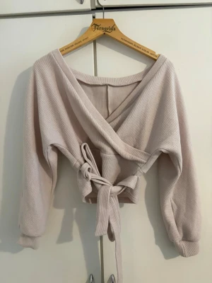 Beige omlottstickad tröja med knytband - Supersnygg beige omlottopp med långa ärmar och breda ribbade muddar. Tröjan har ett knytband i midjan som ger en snygg siluett och är gjord i mjukt, ribbat material. Några tråd har åkt ut men inga hål liksom de kan man se på andra bilden!