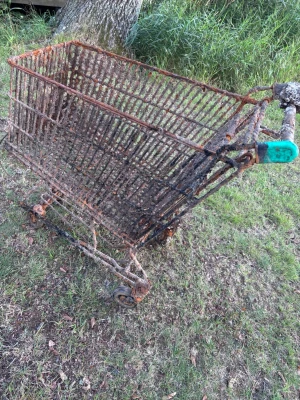 Rostig kundvagn i metall - En gammal kundvagn i metall med tydlig rost över hela ytan. Vagnen har fyra hjul och ett handtag med en grön plastdetalj. Perfekt för dig som vill ha en unik inredningsdetalj eller behöver en vagn till kreativa projekt.