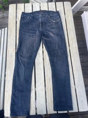 Nudie Jeans Co Mörka Jeans - Mörkblå/otvättade jeans från Nudie Jeans Co. Tillverkade i högkvalitativ denim med fin patination. Gott skick överlag. Notera litet hål på vänster ben. Lätt fixat och nästan osynligt då. Jag kan göra det eller du. Det är även litet hål i vänster ficka, också lätt fixat, syns inte utifrån. 